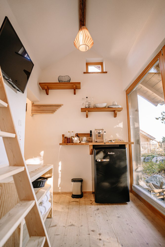 Innenansicht eines Tiny Houses am Mächlerhof mit heller Küchenzeile, offenem Regal, kleinem Kühlschrank und großem Fenster mit Blick ins Freie.
